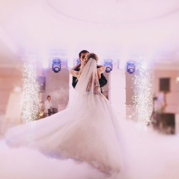 A wedding couple dances in a hall with twinkle lights and a fog effect while a wedding DJ plays music.