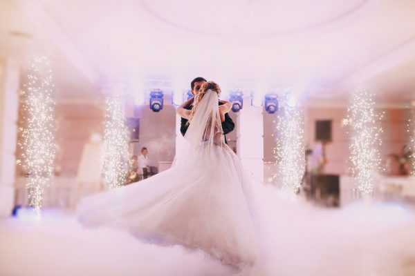 A wedding couple dances in a hall with twinkle lights and a fog effect while a wedding DJ plays music.