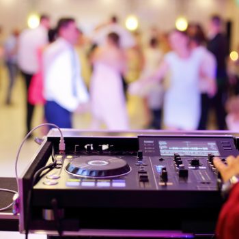 A hand rests on the turntable as a wedding DJ plays music with people dancing in the background.