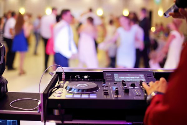 A hand rests on the turntable as a wedding DJ plays music with people dancing in the background.
