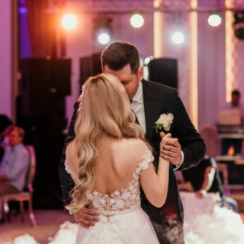 A bride with long blonde hair dances with a tall groom in a suit, with warm-colored lights, and wedding guests sitting in the background.