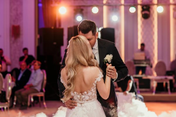 A bride with long blonde hair dances with a tall groom in a suit, with warm-colored lights, and wedding guests sitting in the background.