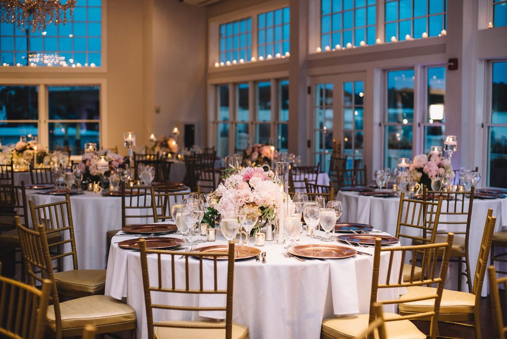 Tables and chairs decorated for a wedding reception in a seaside ballroom with wall-to-wall windows.