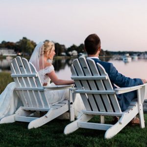 A bride and groom sit in white Adirondack chairs overlooking a harbor at dusk.