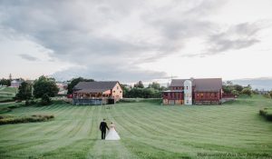 A groom and bride walk up a green lawn toward a barn and restaurant at a wedding venue in MA.