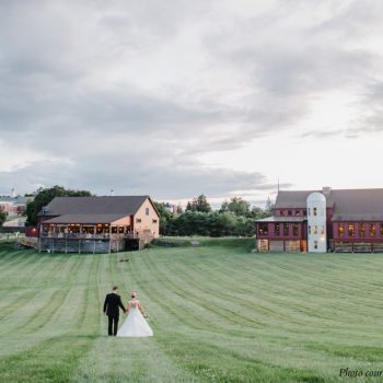 A groom and bride walk up a green lawn toward a barn and restaurant at a wedding venue in MA.