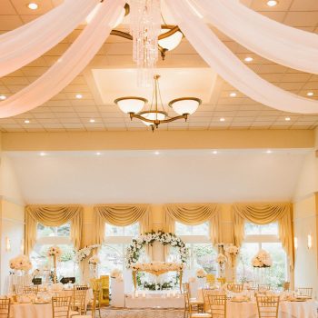 A ballroom glows with warm light, with white fabric and chandeliers hanging from a high ceiling at a wedding venue in NH.