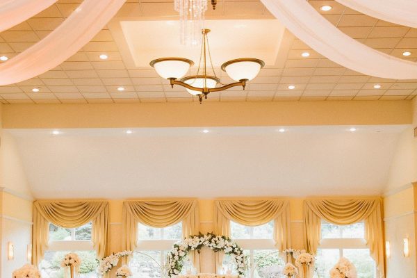 A ballroom glows with warm light, with white fabric and chandeliers hanging from a high ceiling at a wedding venue in NH.
