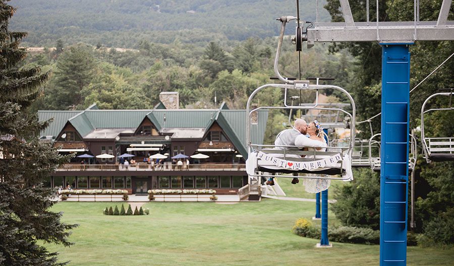 A bride and groom sit on a chairlift, with a mountain lodge in the background, at one of the ski resort wedding venues in NH.