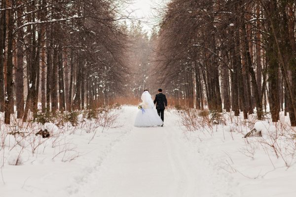 A bride and groom walk along a snowy path flanked by trees during a winter wedding scene.