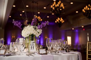 A table at a wedding, with florals and glassware, with amber-colored chandeliers, and purple DJ Uplighting along the walls.