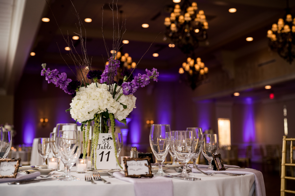 A table at a wedding, with florals and glassware, with amber-colored chandeliers, and purple DJ Uplighting along the walls. 