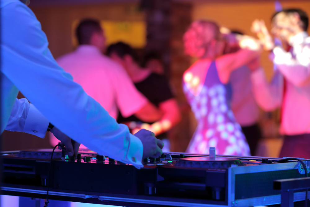 A DJ's hands rest on the sound board while guests dance under colored lights at an event