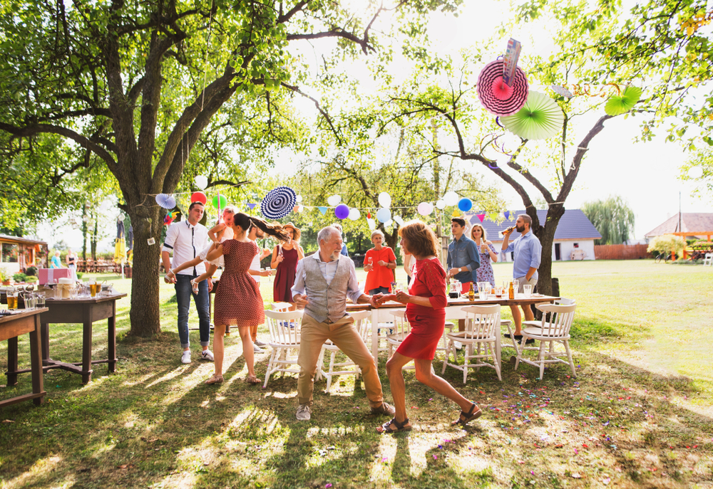 An older couple and other party guests dance in front of a picnic table outside on a sunny day