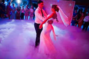 A groom and bride dance in the center of the dance floor with pink lights and cloud-like mist.