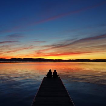 A couple sits at the end of a dock on Lake Champlain watching the sun set behind mountains