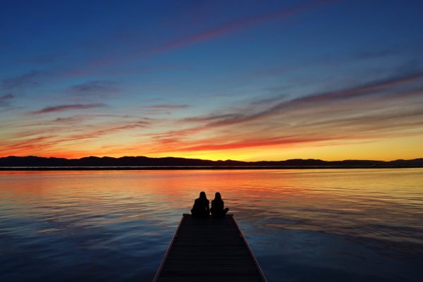 A couple sits at the end of a dock on Lake Champlain watching the sun set behind mountains