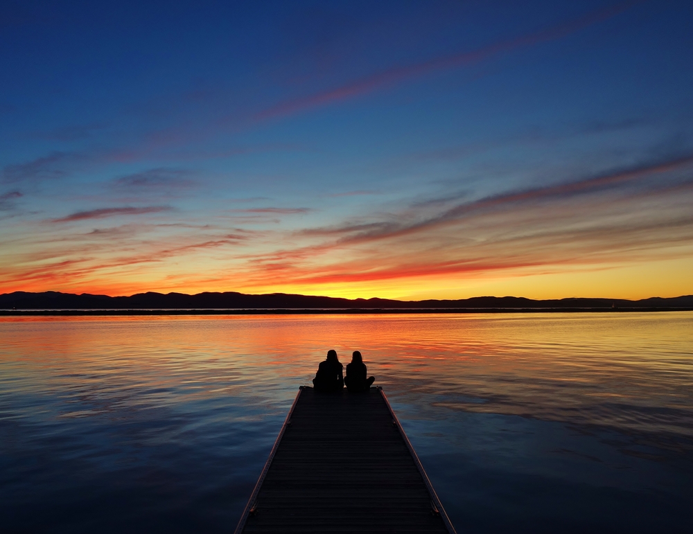 A couple sits at the end of a dock on Lake Champlain watching the sun set behind mountains