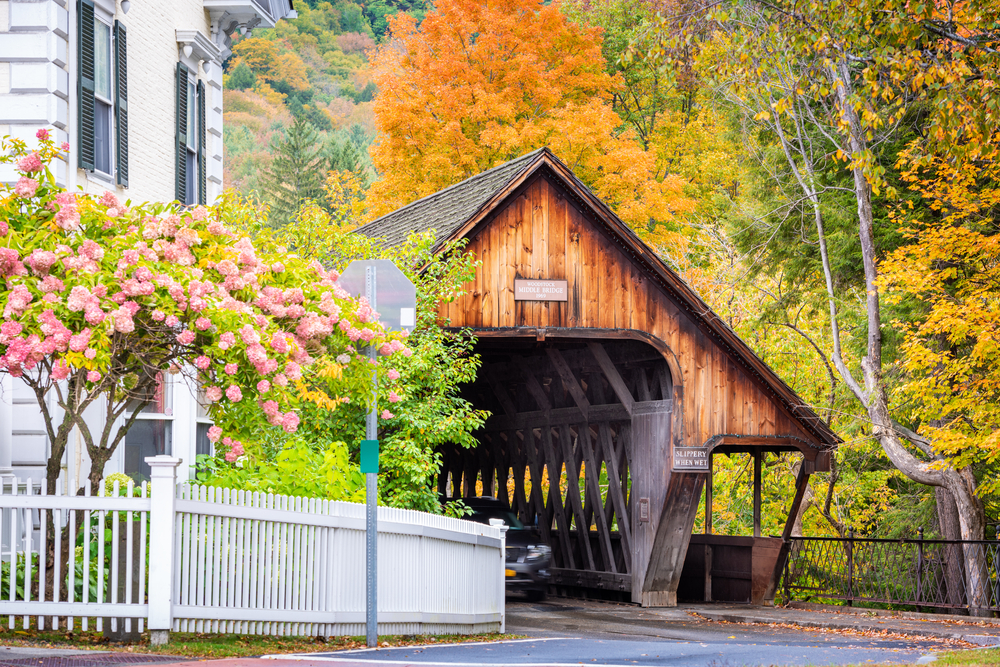 A wooden covered bridge in Vermont with yellow fall foliage behind. 