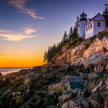 Bass Head Lighthouse sits on a rocky cliff while the sun sets behind the water.
