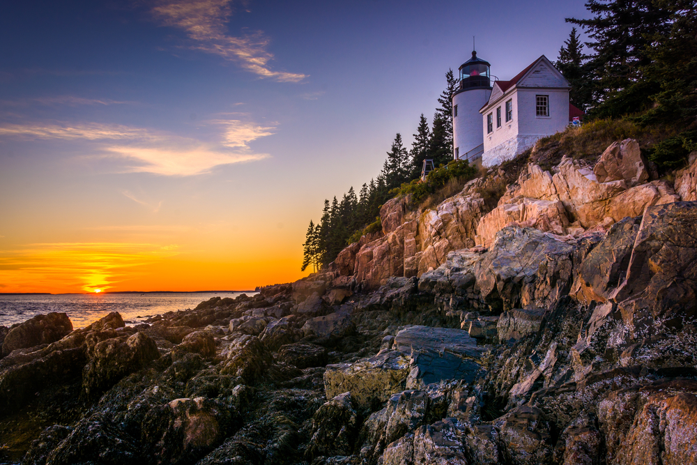 Bass Head Lighthouse sits on a rocky cliff while the sun sets behind the water.