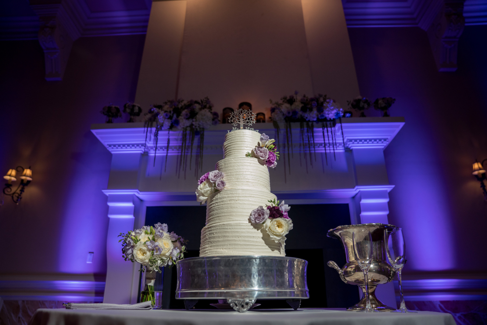 A wedding cake on a pedestal with two purple DJ UpLights illuminating the large fireplace behind.