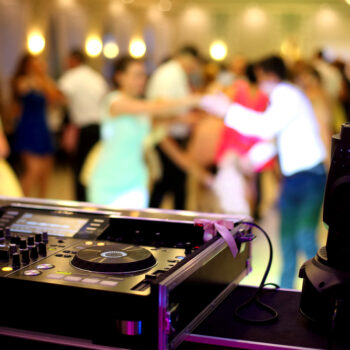 A DJ mixer sits in the foreground while a group of people dance during a wedding.
