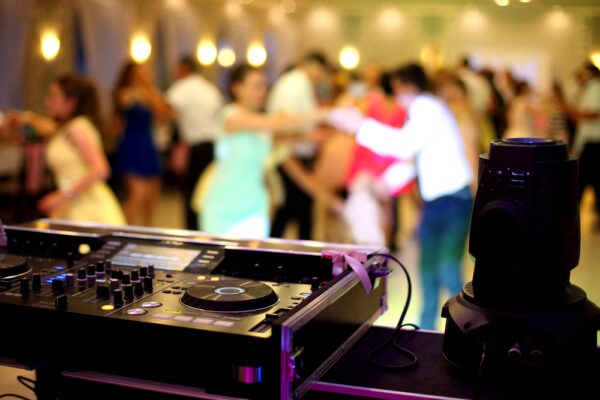 A DJ mixer sits in the foreground while a group of people dance during a wedding.