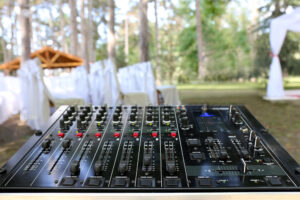 A dj mixer set up at an outdoor wedding, with white fabric draping chairs and tall trees in the background.